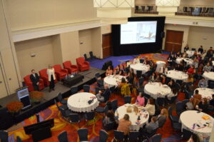 A photo from a hotel ballroom mezzanine with two people on a stage speaking to a large group of people seated around multiple circular tables.